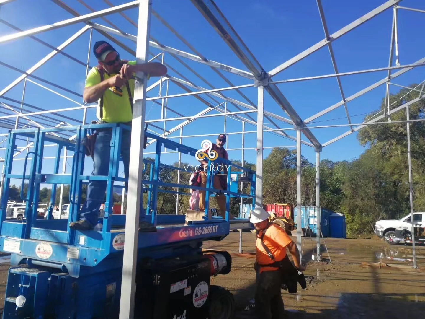 Workers installing greenhouse steel framework from a raised platform, with another worker at ground level
